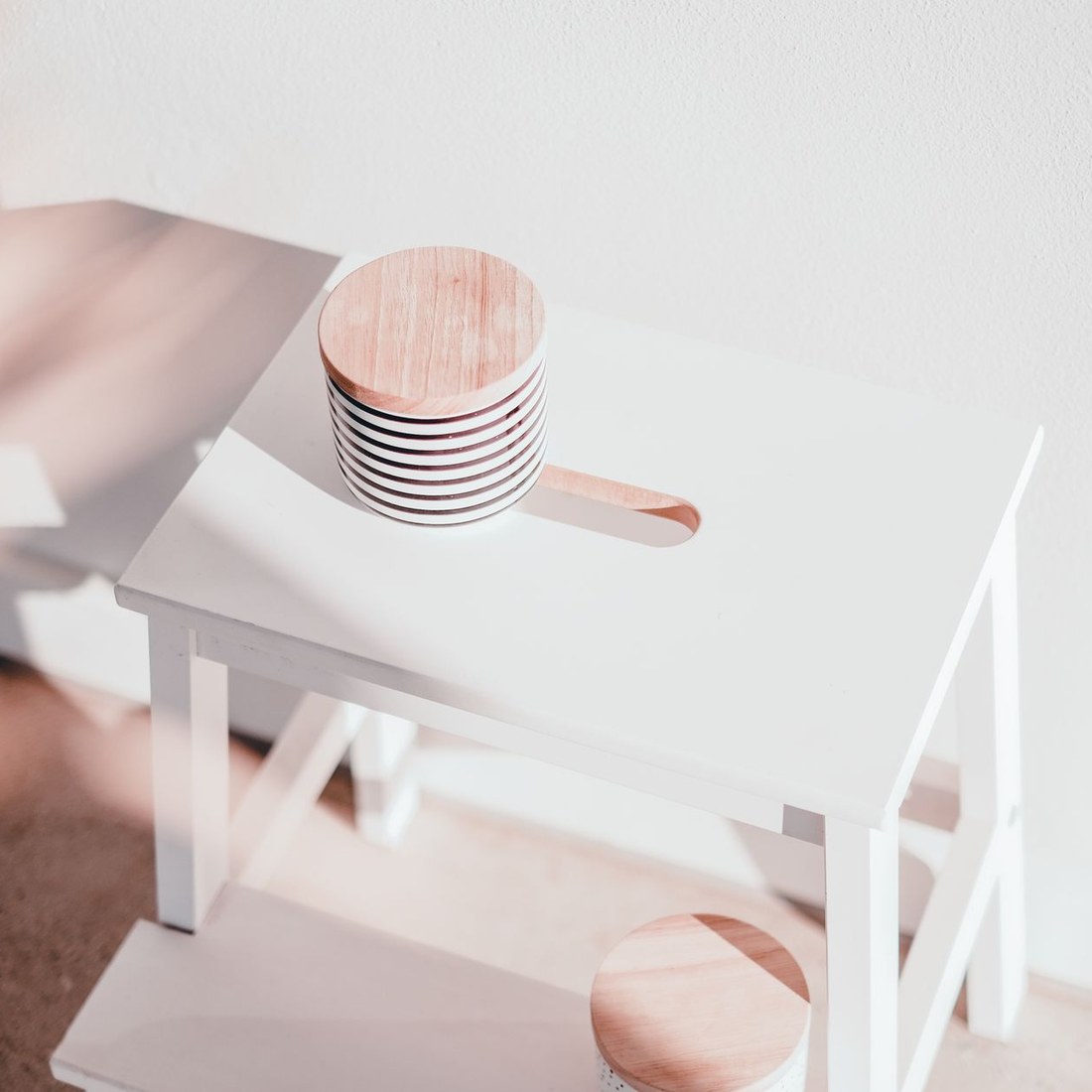 A child's white wooden step stool in bright natural light against a white wall, with stacked wooden coasters resting on the seat
