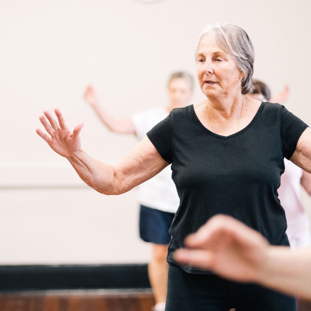 Older woman moving her arms through a slow exercise in a bright community class, with other participants behind her
