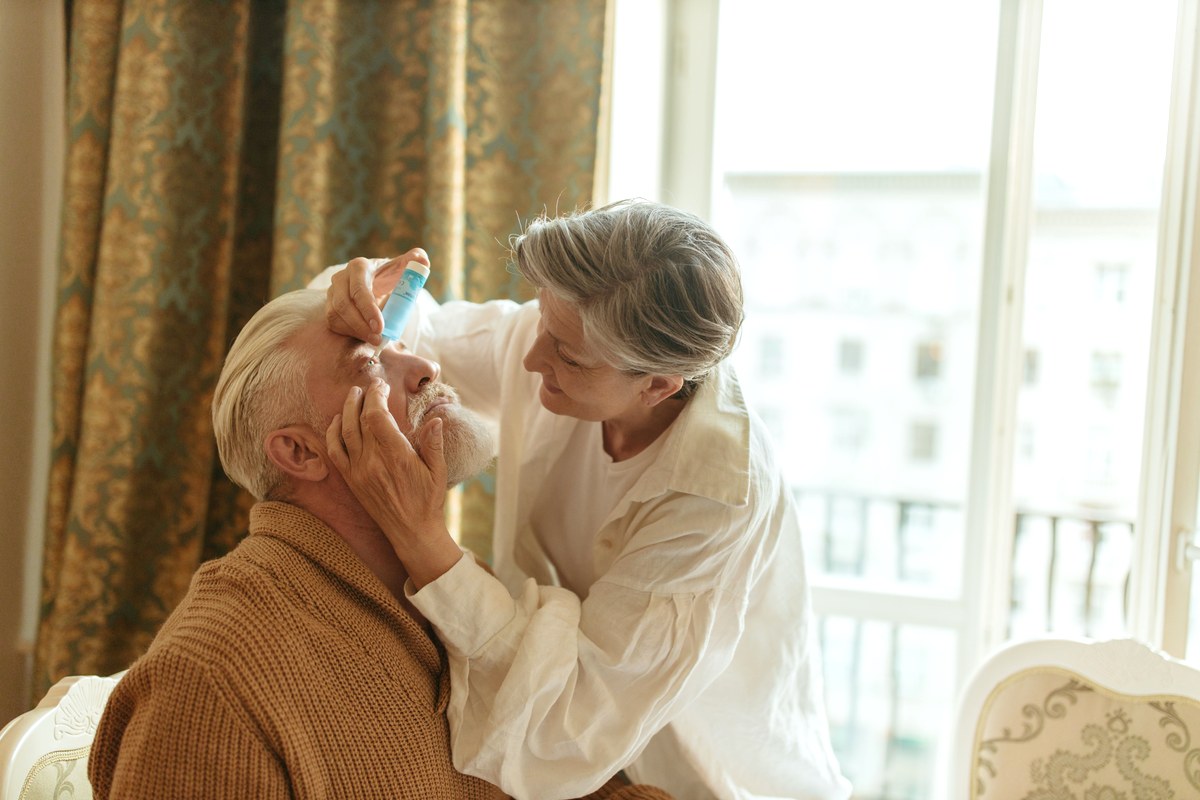 An older woman steadying her partner's face while putting an eye drop in for him beside a sunlit window