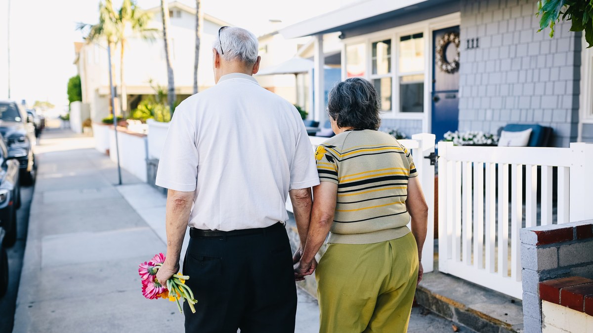 An older couple holding hands and walking away along a sunlit residential sidewalk, one of them carrying a small bunch of flowers.