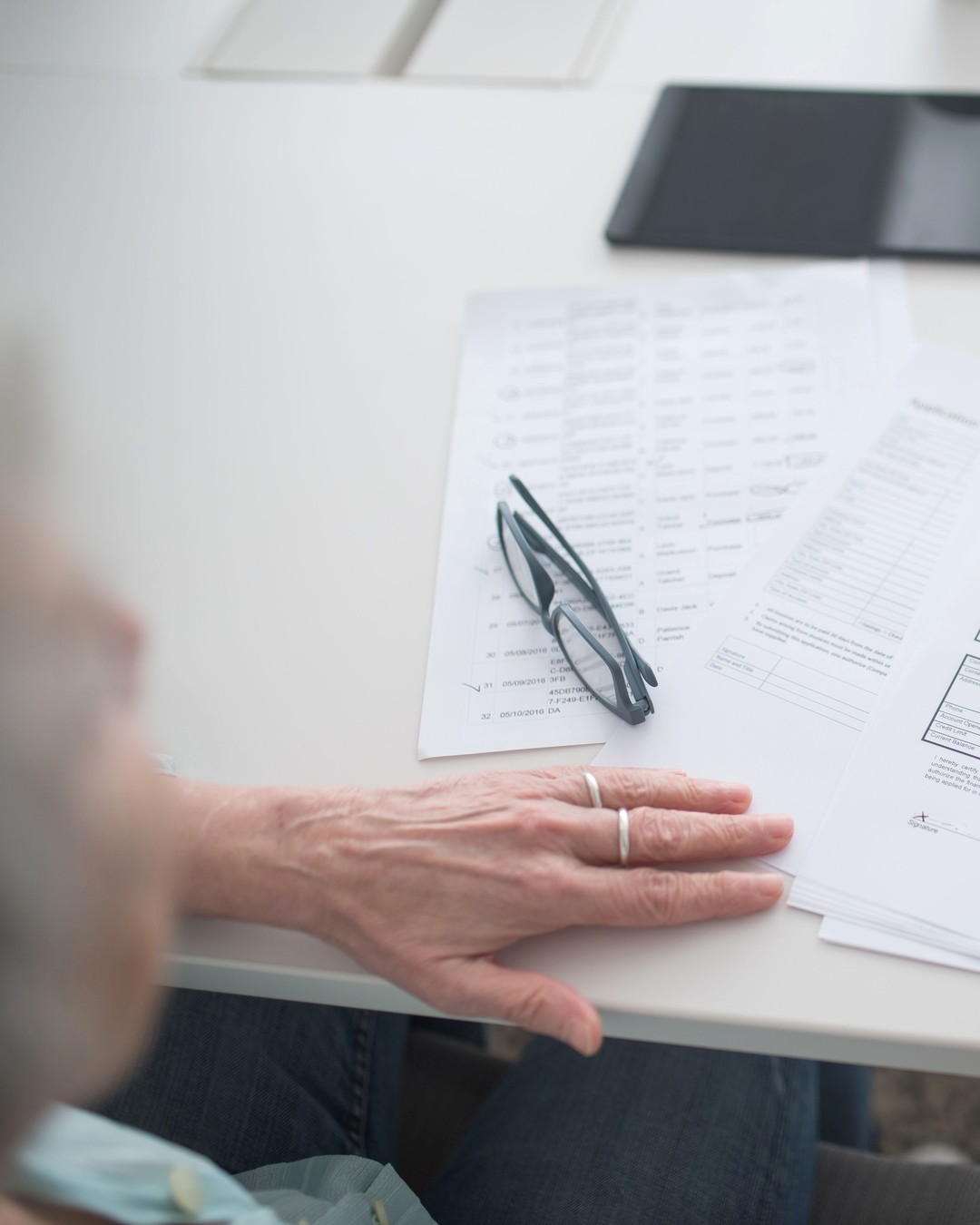 An older person's hand resting on a printed sheet of results at a bright white table, reading glasses folded on top of the page, face not visible