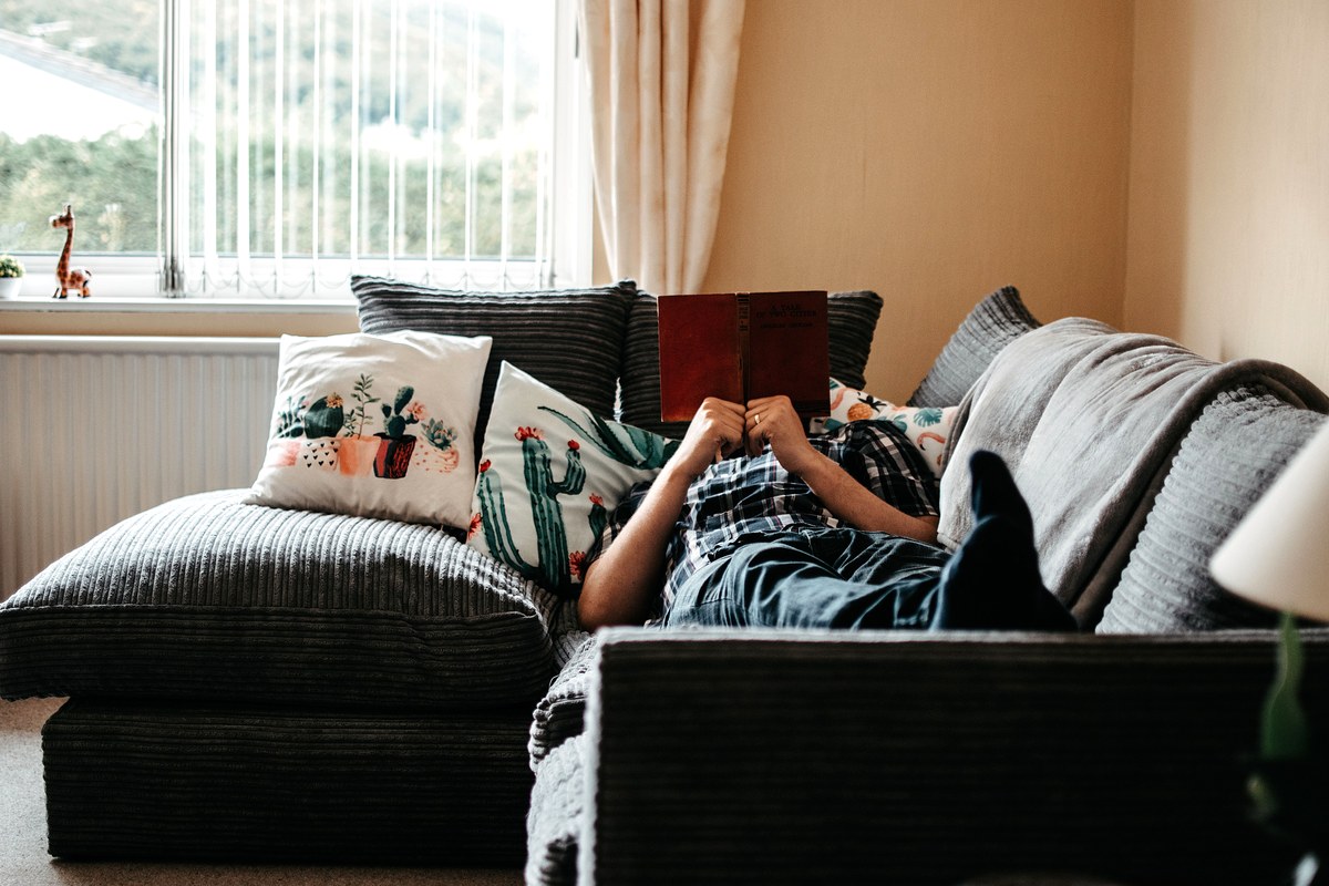A person lying on a living room couch during the day with their legs propped up on the cushions, reading