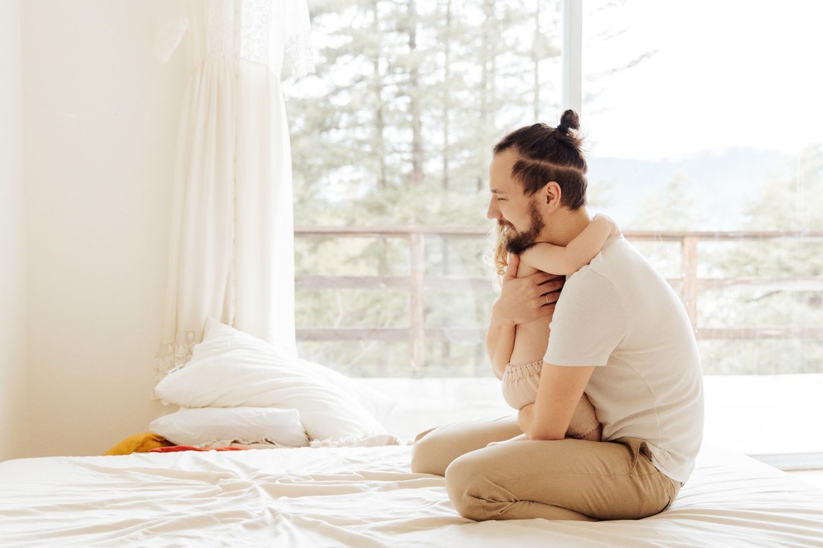A father sitting on a bed by a sunlit window, hugging his young child