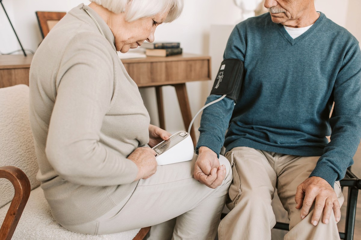An older woman fitting an upper-arm cuff and holding a digital blood pressure monitor for an older man at home