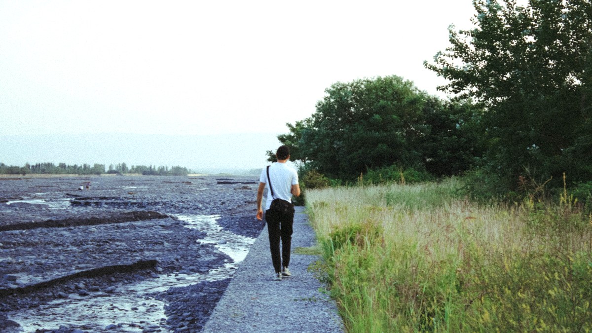 A man walking away along a riverside path lined with grass and trees in the late afternoon