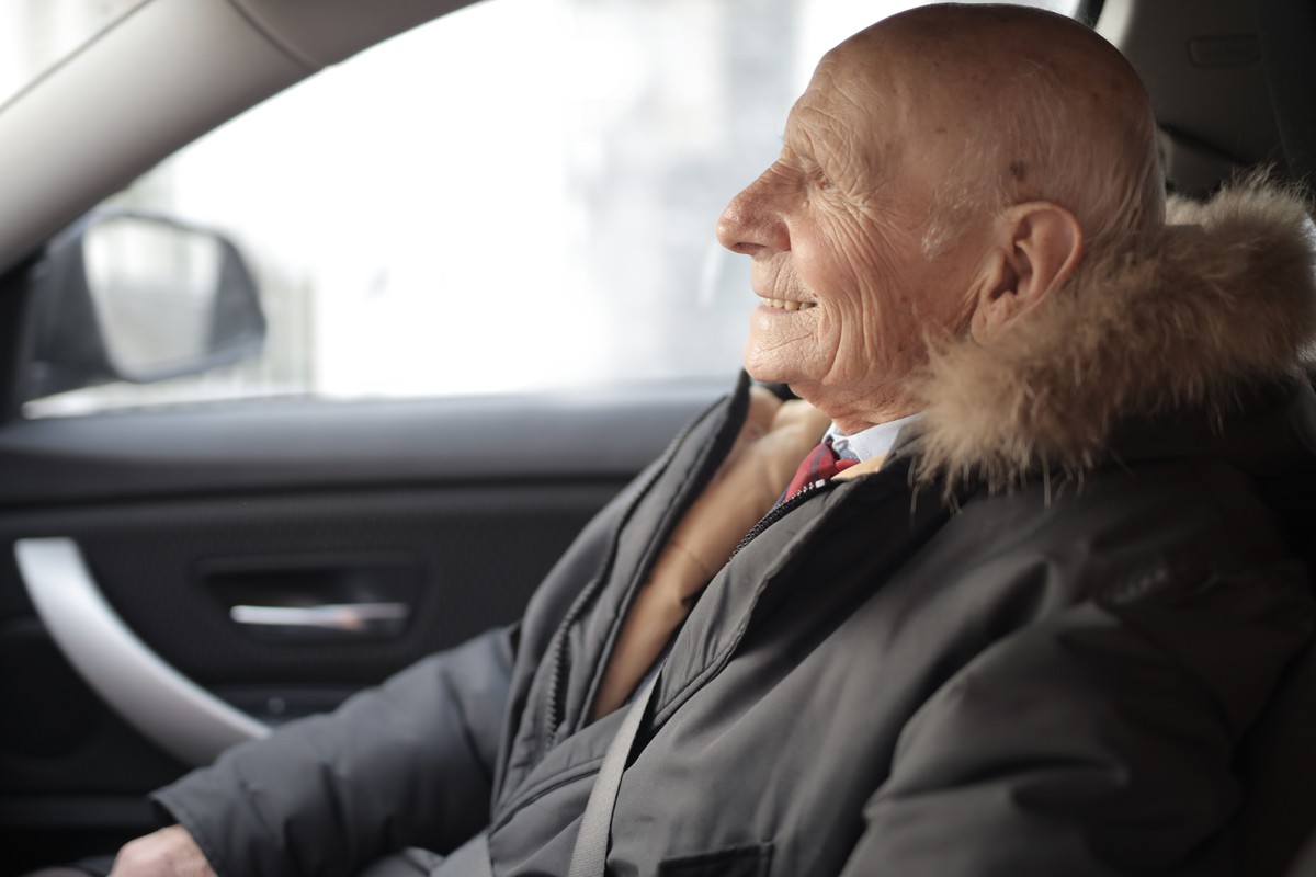 A smiling older man sitting in the front passenger seat of a car with the seat belt across his coat