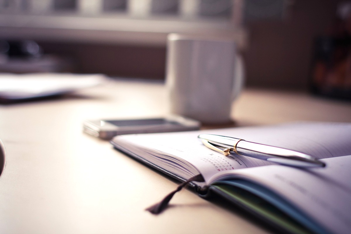 An open paper planner with a pen resting across the page, a white mug and a phone with a blank screen on a desk in soft daylight
