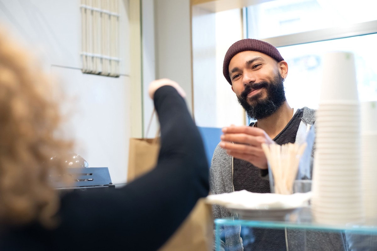 A café worker smiling at a customer across the counter, beside a paper bag and a stack of takeaway cups in daylight