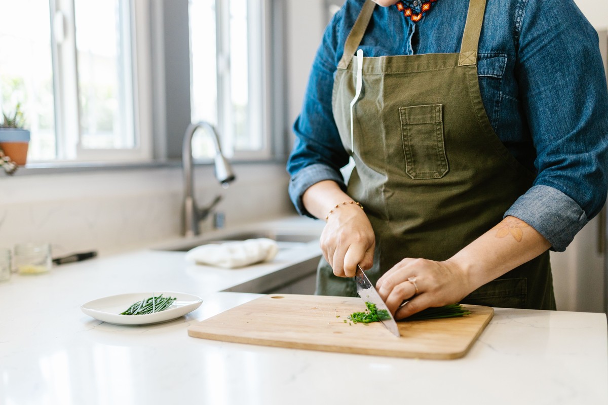 A home cook in an apron chopping chives with a chef's knife on a wooden cutting board in a bright kitchen