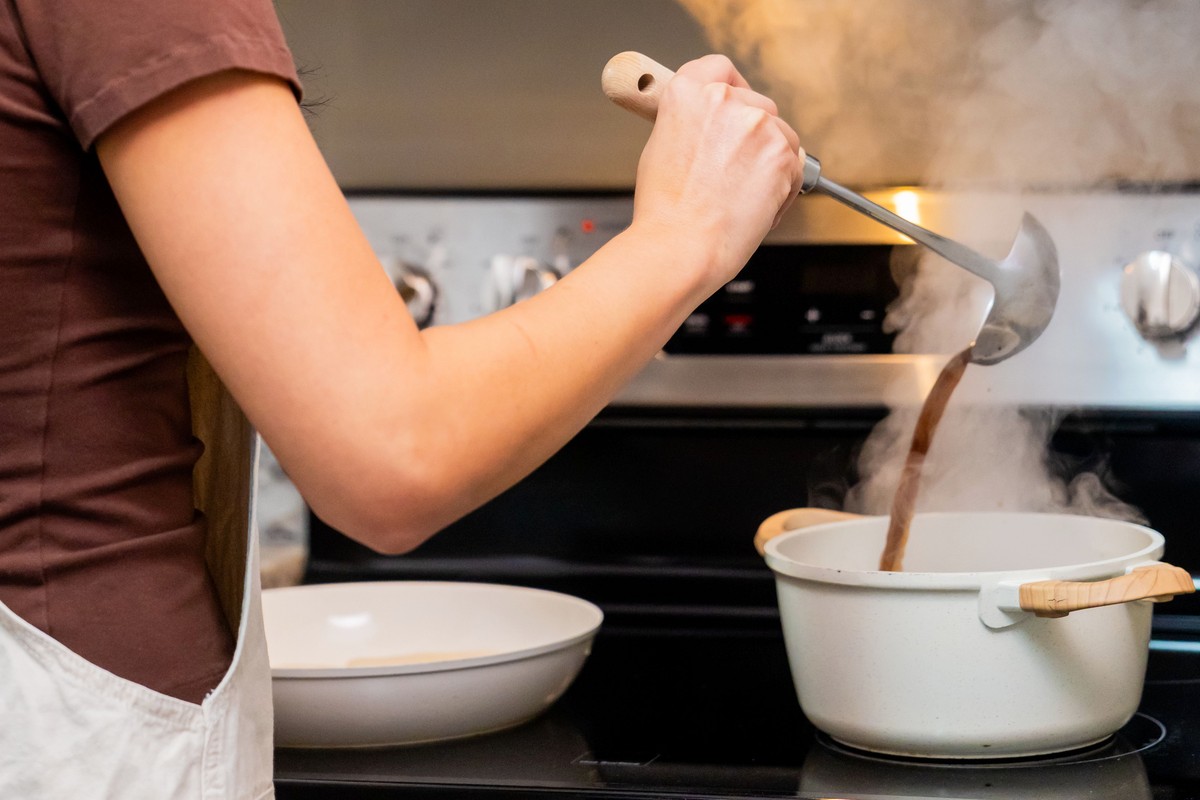 A home cook in an apron ladling steaming stew from a large white pot on the stove into a shallow bowl