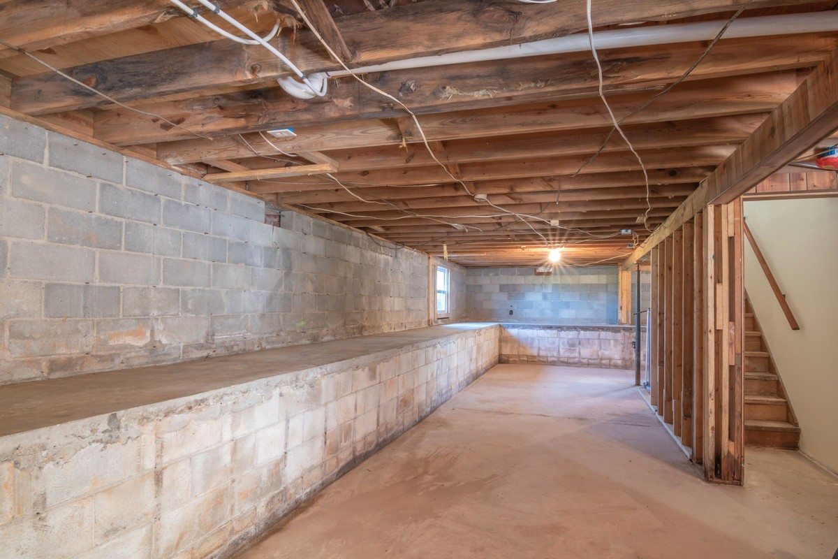 An unfinished basement with concrete block walls, a poured slab and exposed floor joists - the lowest lived-in level of a house, where soil gas arrives first and where a radon test kit belongs
