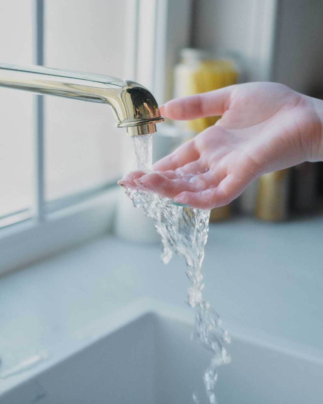 An open hand held under water running from a brass kitchen tap beside a bright window, over a white sink