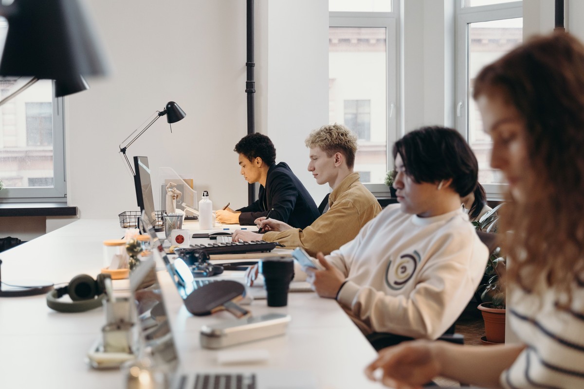 Office workers sitting side by side at a long shared desk with laptops, a keyboard and a desk lamp near bright windows