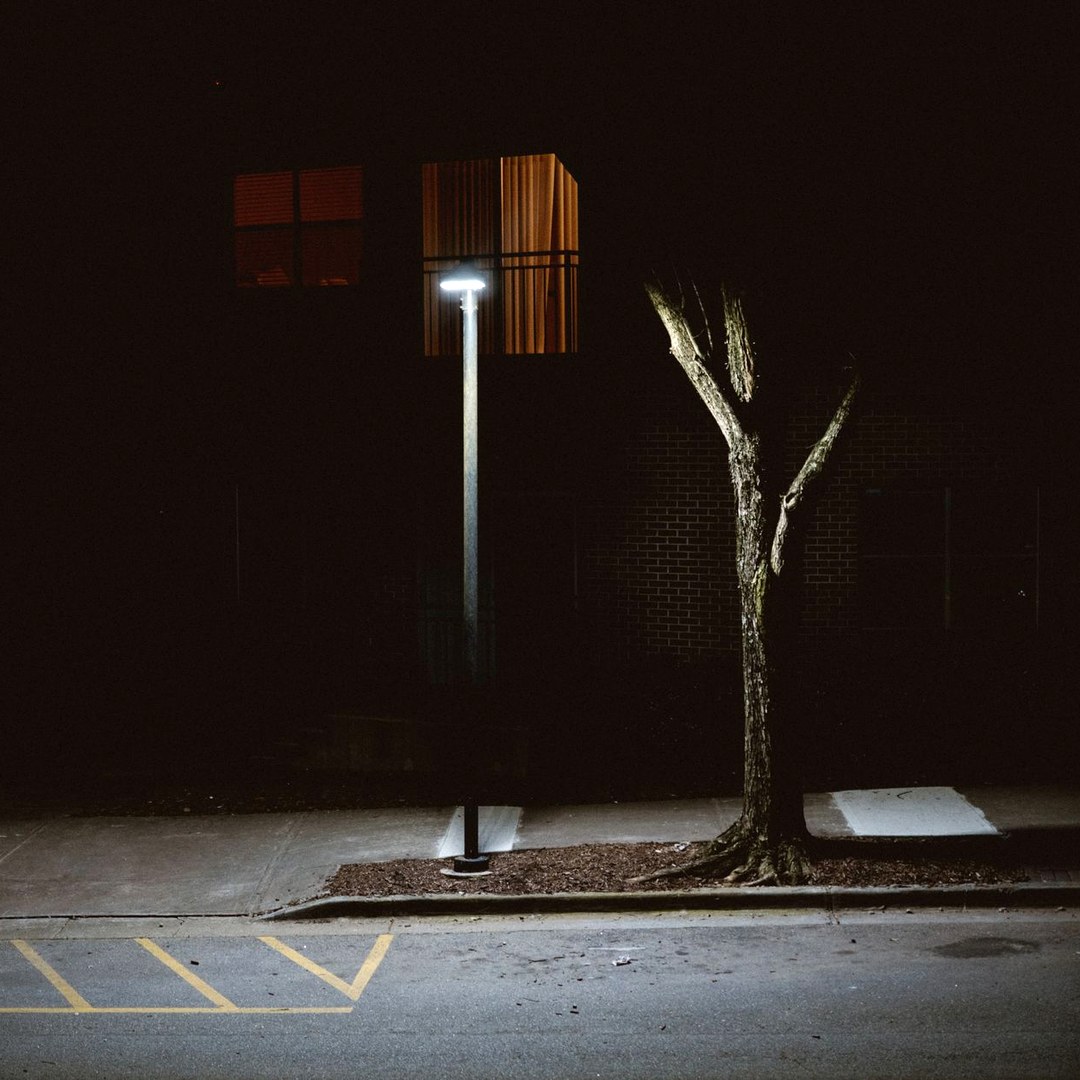 An empty street at night lit by a single lamp post, with one curtained window still glowing in the building behind it