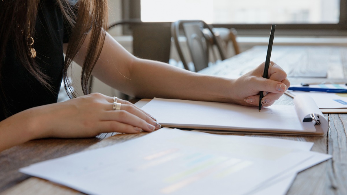 Close-up of a person’s hands writing on a pad of paper at a wooden table beside a bright window