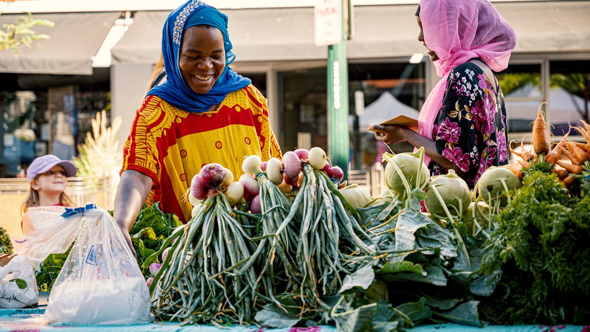A market vendor and a shopper smiling at each other across a stall of fresh vegetables at a sunny farmers market
