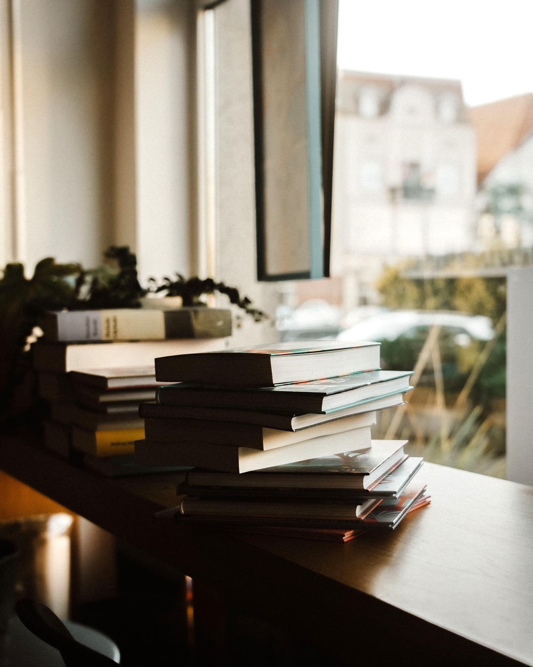 A stack of hardback and paperback books resting on a wooden ledge beside a bright window