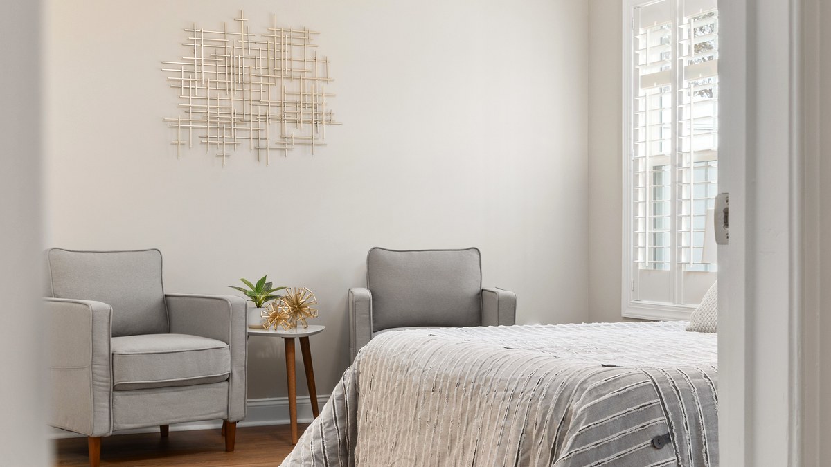 A bright bedroom with two sturdy armchairs and a small side table beside the bed, a clear wooden floor and daylight through the shutters