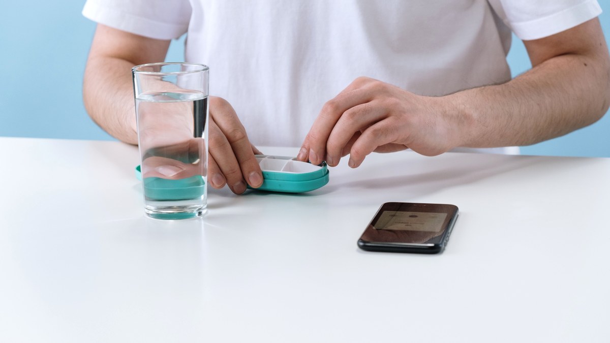A person at a plain white table opening a weekly pill case beside a glass of water and a smartphone