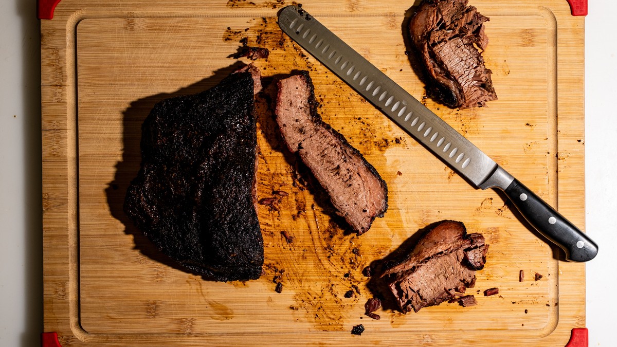 A cooked beef brisket part-sliced into portions on a bamboo cutting board with a long slicing knife, seen from directly above