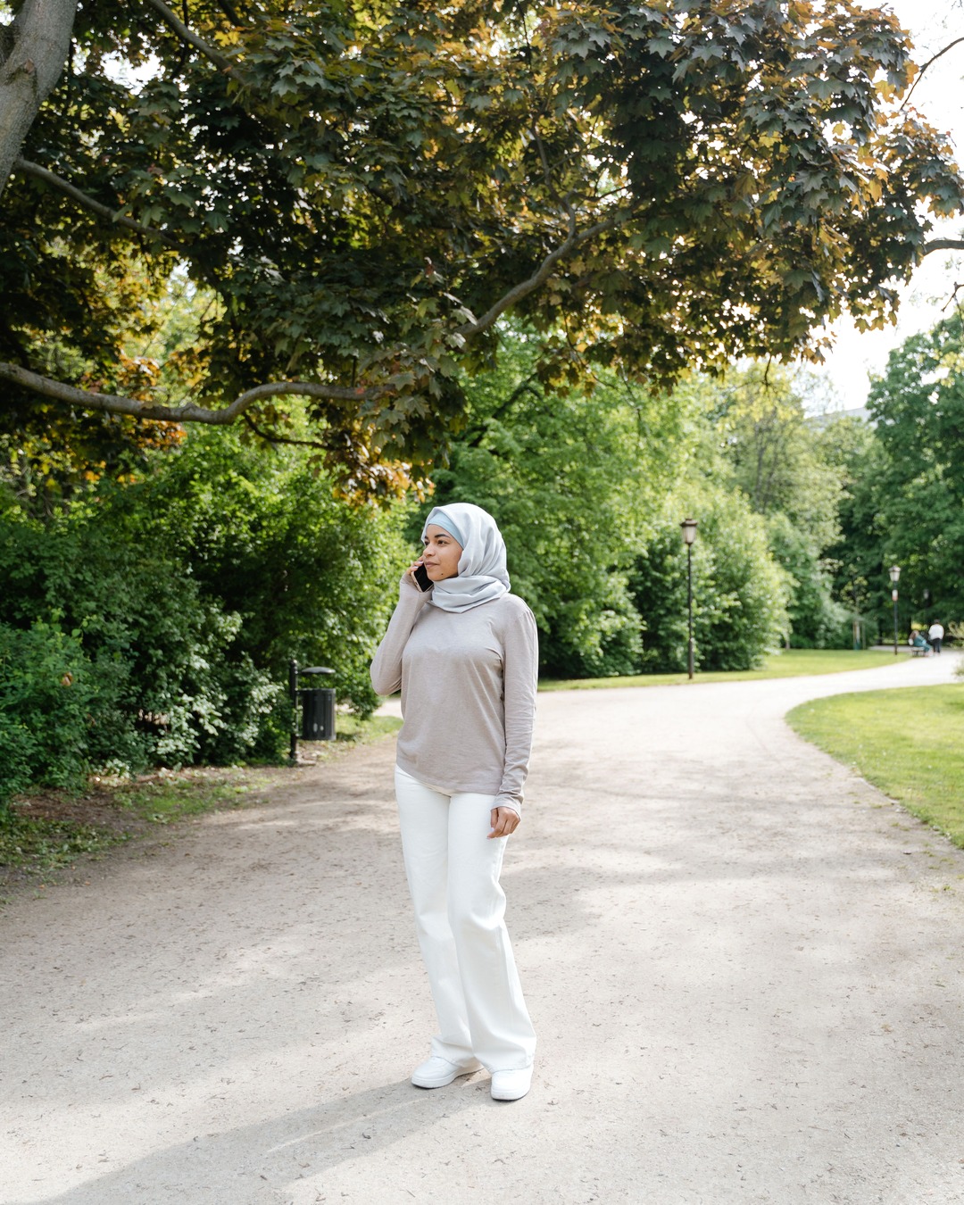 A woman standing on a tree-lined gravel path in a park on a bright day, talking on her phone