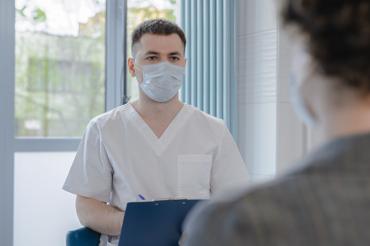 A clinician in scrubs holding a clipboard listens to a patient during a consultation in a bright clinic room