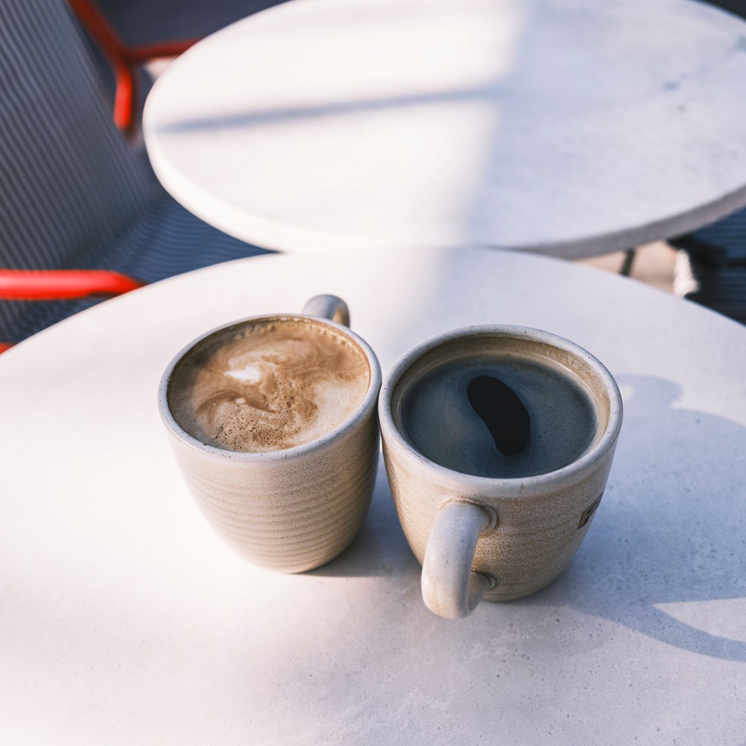 Two coffee cups standing side by side on a sunlit café table, with an empty table and chair behind them
