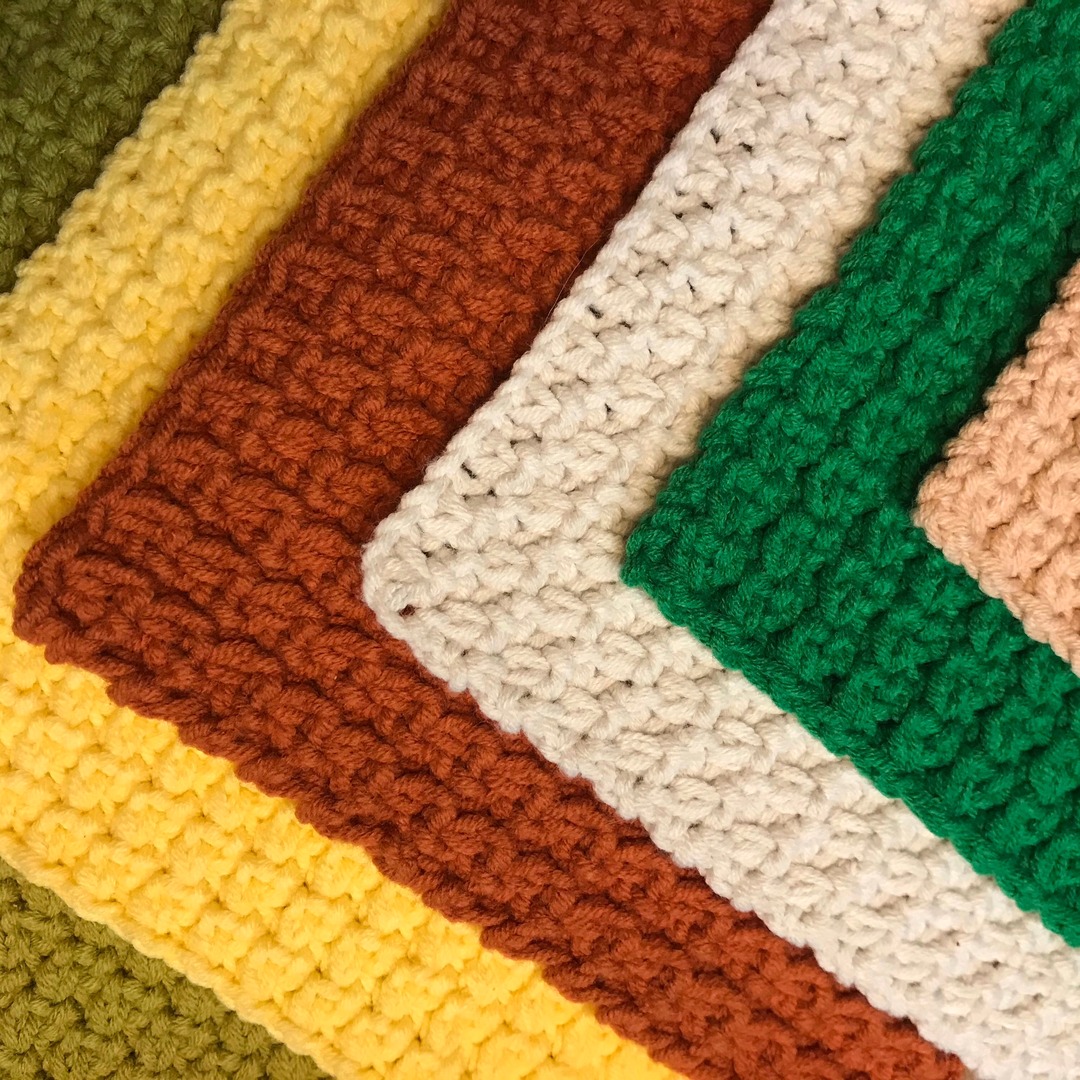 Close-up of several crocheted wool squares in olive, yellow, rust, cream and green, photographed in flat daylight so the stitch texture is clearly visible