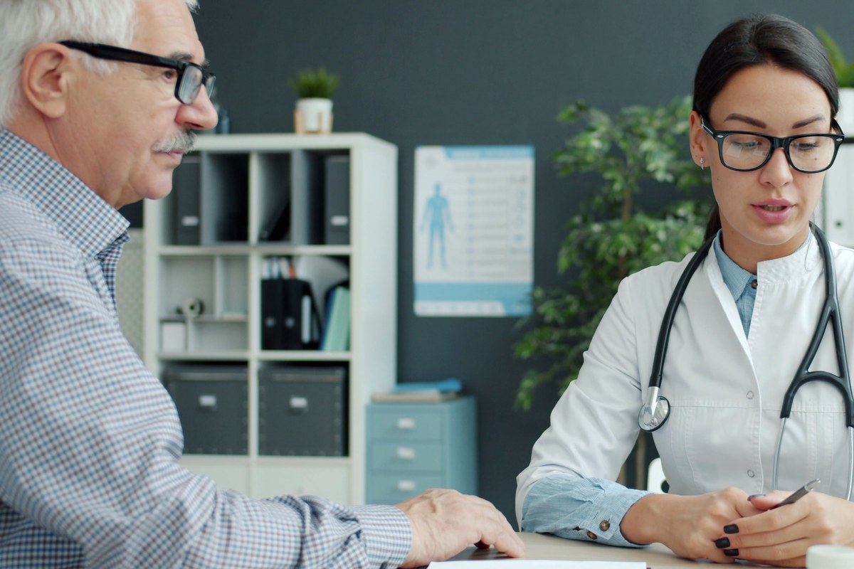 An older man talking with a clinician across a desk in a bright medical office