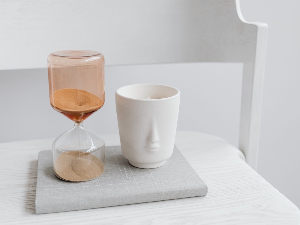 A glass hourglass with amber sand beside a white ceramic cup on a grey book in a bright, airy room
