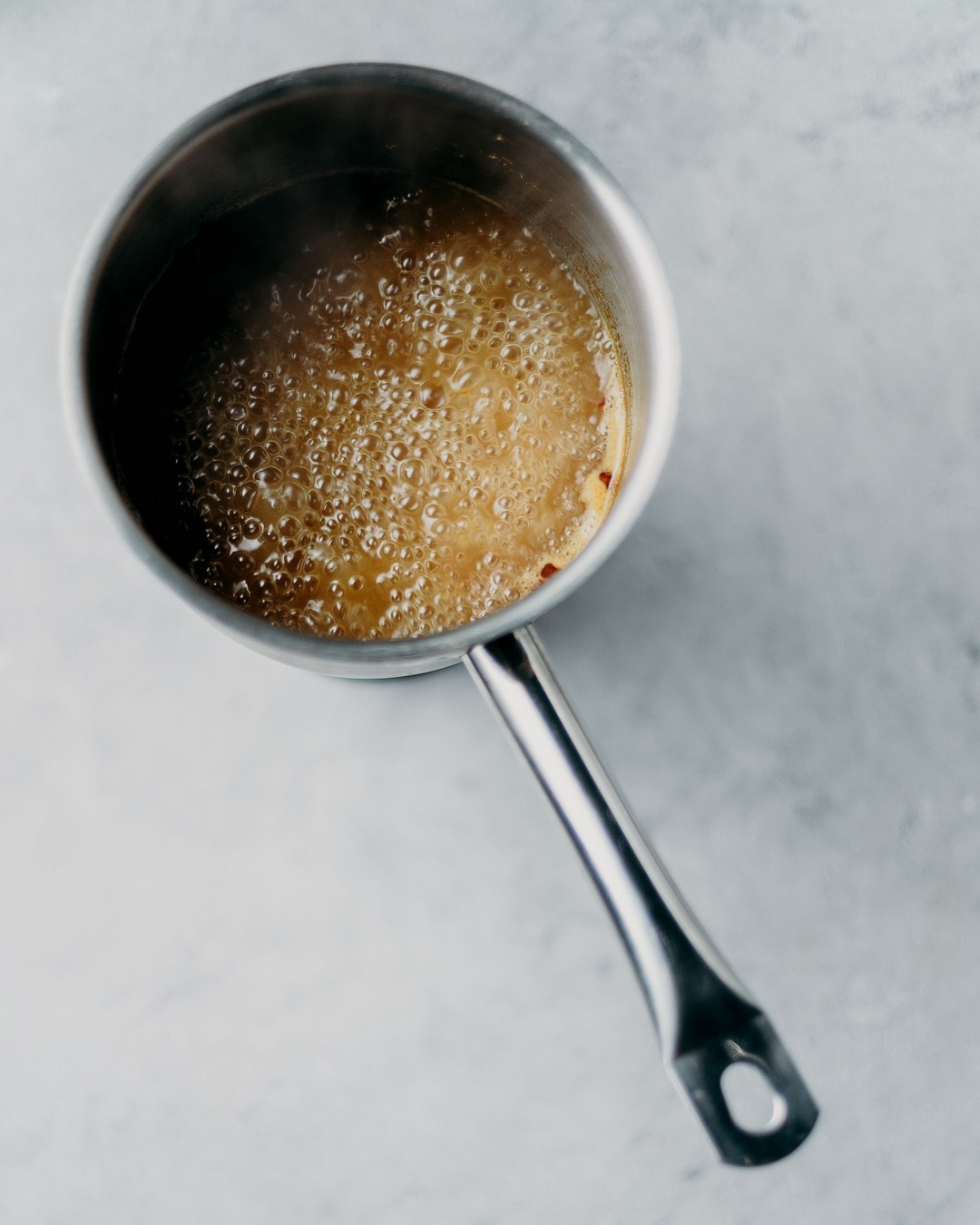 A stainless steel saucepan of amber broth at a full rolling boil, seen from directly above on a pale stone counter
