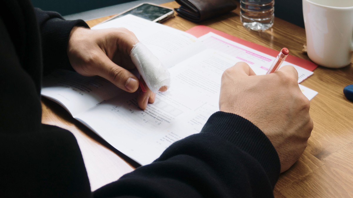 A man writing at a wooden desk with a gauze-wrapped index finger, going about his day while the cut heals
