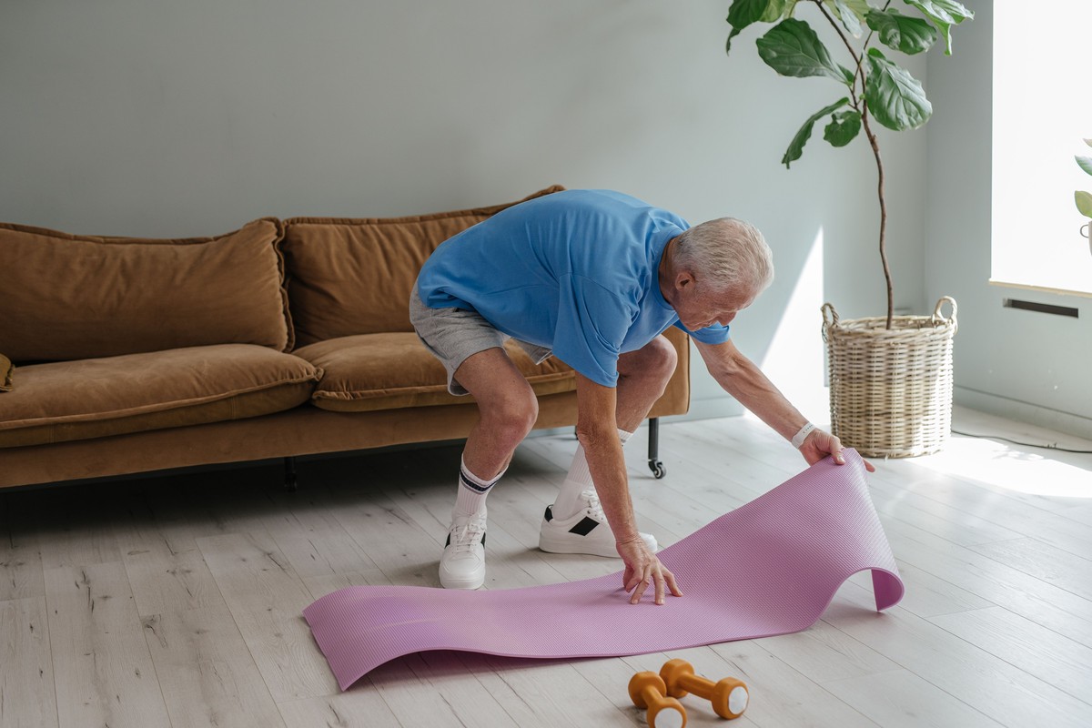 An older man in a blue T-shirt unrolling an exercise mat on his living room floor, with a pair of small dumbbells beside it