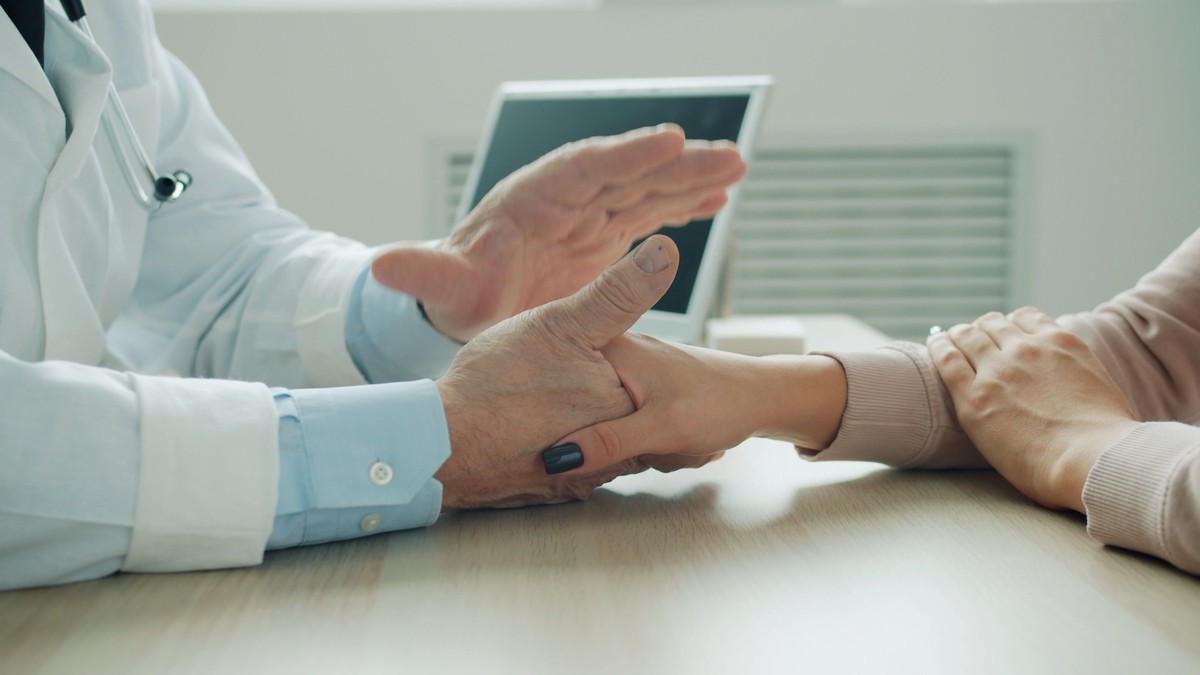 A doctor holds a patient's hand and wrist across a desk during an exam in a bright medical office